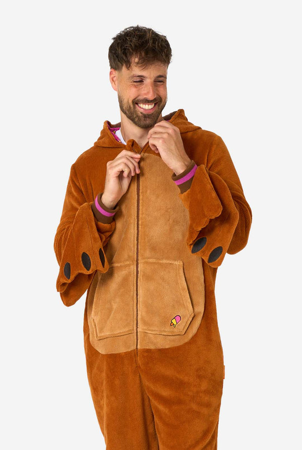 Ein fröhlicher Mann trägt einen braunen Erwachsenen-Onesie in Walross-Optik. Der Onesie hat eine Kapuze mit Reißzähnen, aufgenähte Handschuhe in Pfotenform und verspielte Walross-Details – perfekt zum gemütlichen Faulenzen oder für Kostümpartys. Die Kapuze hängt ihm über den Rücken.