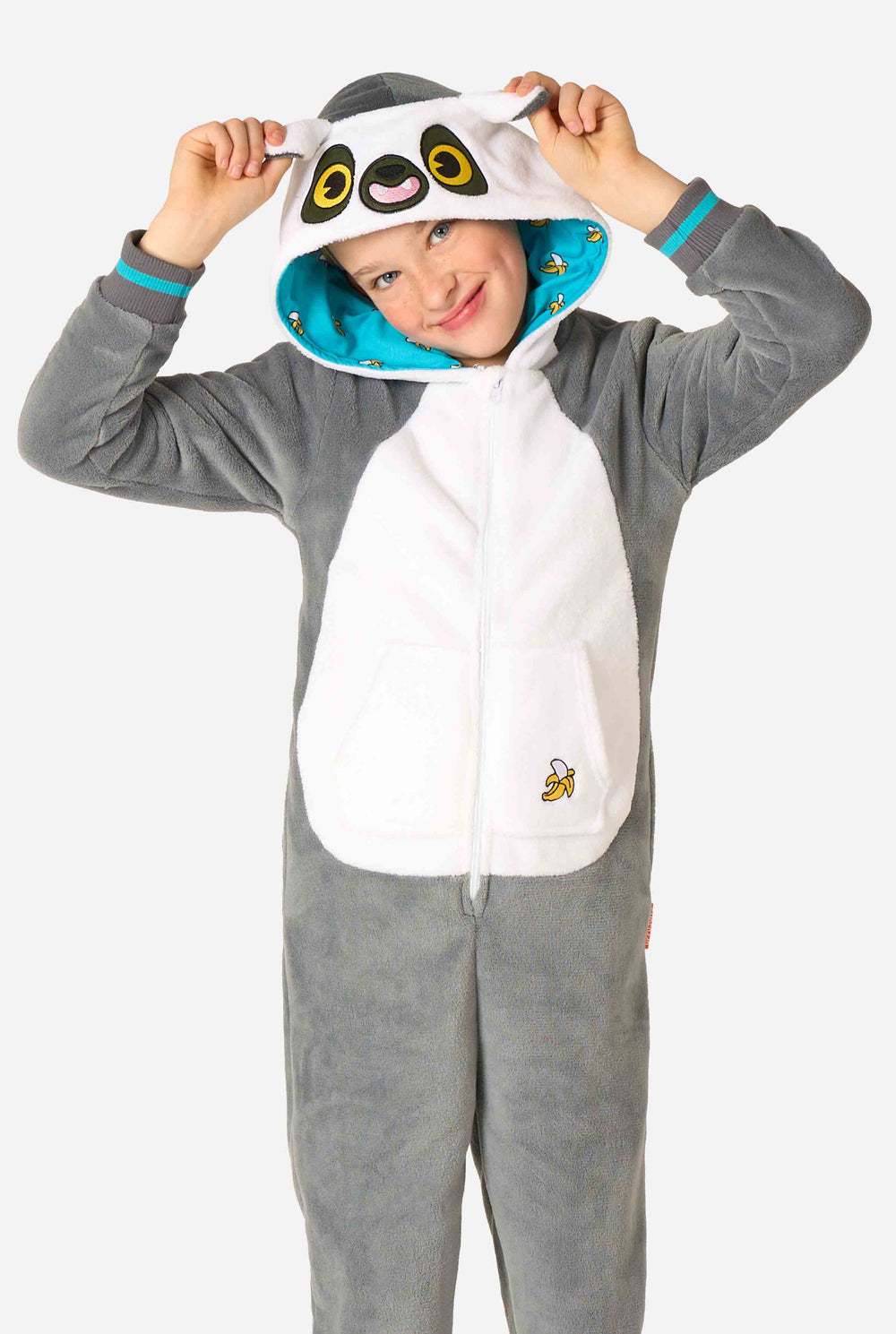 Kind trägt einen gemütlichen, unisex Kinder-Onesie im Lemur-Design aus grau-weißem Plüschstoff. Die Kapuze zeigt ein Cartoon-Lemurgesicht mit Ohren, und auf der Tasche befindet sich ein verspielter Bananenaufdruck.