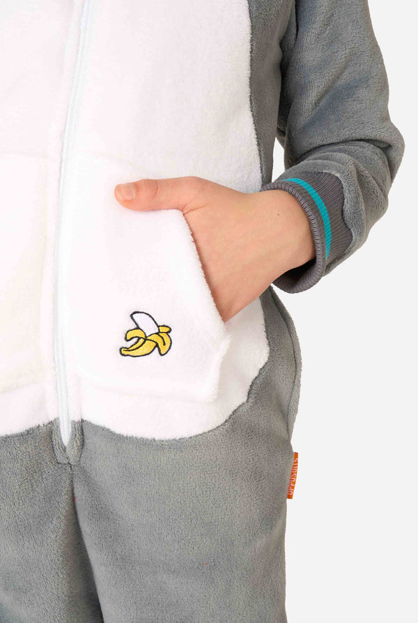 Kind trägt einen gemütlichen, unisex Kinder-Onesie im Lemur-Design aus grau-weißem Plüschstoff. Die Kapuze zeigt ein Cartoon-Lemurgesicht mit Ohren, und auf der Tasche befindet sich ein verspielter Bananenaufdruck. Fokus auf eine Hand in der Tasche.