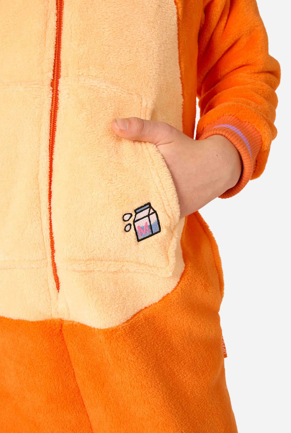 Kind trägt einen verspielten, unisex Kinder-Onesie im Design einer rot-orangefarbenen Katze, mit Kapuze, die ein Cartoon-Katzengesicht und Ohren zeigt, sowie einem hellbeigen Bauchpaneel. Das Kind zieht eine lustige Grimasse vor einem schlichten Hintergrund. Fokus auf eine Hand in der Tasche.