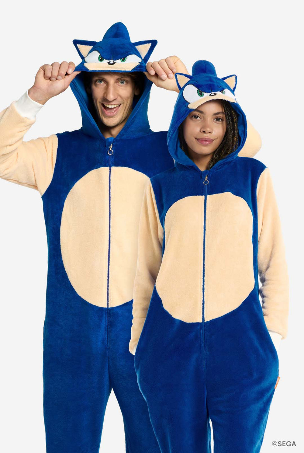 Ein Mann und eine Frau tragen einen verspielten blauen und beigen Sonic the Hedgehog-Strampler für Erwachsene von OppoSuits. Der kuschelige Strampler hat einen Reißverschluss auf der Vorderseite und eine angesetzte Kapuze, die Sonics ikonischem Gesicht nachempfunden ist, komplett mit Ohren und einem ausdrucksstarken Blick. Der blaue Körper des Anzugs kontrastiert mit einem beigen Bauch, der den klassischen Look von Sonic nachahmt. 