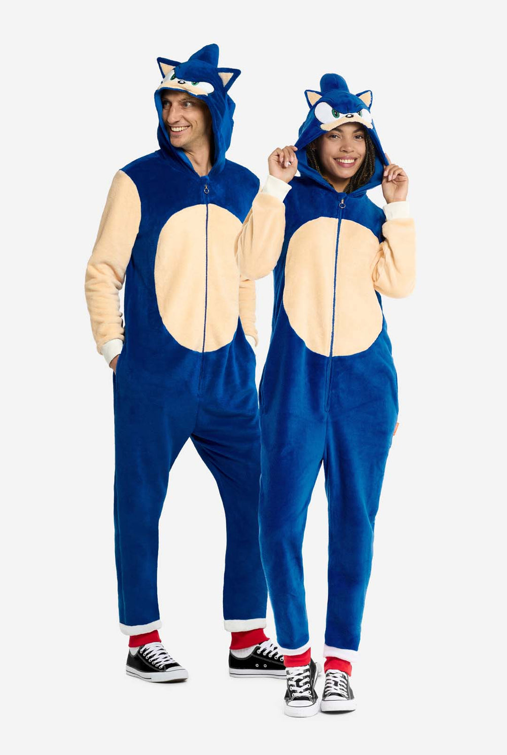 Ein Mann und eine Frau tragen einen verspielten blauen und beigen Sonic the Hedgehog-Strampler für Erwachsene von OppoSuits. Der kuschelige Strampler hat einen Reißverschluss auf der Vorderseite und eine angesetzte Kapuze, die Sonics ikonischem Gesicht nachempfunden ist, komplett mit Ohren und einem ausdrucksstarken Blick. Der blaue Körper des Anzugs kontrastiert mit einem beigen Bauch, der den klassischen Look von Sonic nachahmt. 