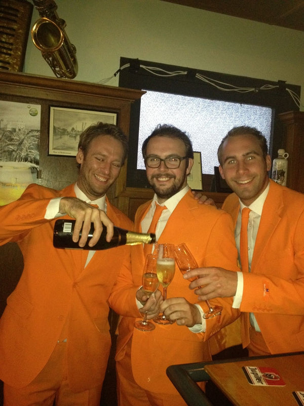 Ein Bild der drei Gründer von Opposuits, die einen orangefarbenen Anzug tragen und Champagner trinken.